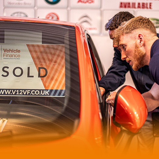 Two people looking to an orange car which has a sold sign in the window